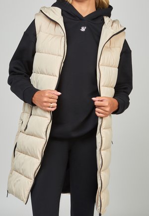 Femme portant une longue doudoune beige par-dessus un sweat à capuche noir et des leggings noirs, tenant la doudoune ouverte avec les deux mains.