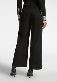 Pantalones de pierna ancha negros con una textura suave, que presentan una cintura alta y dos bolsillos traseros. Combinados con zapatos de tacón alto negros.