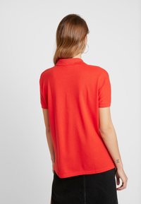 Femme aux cheveux châtain clair, portant de grandes boucles d'oreilles créoles, une polo à manches courtes rouge vif et une jupe noire, tournant le dos à l'appareil photo.