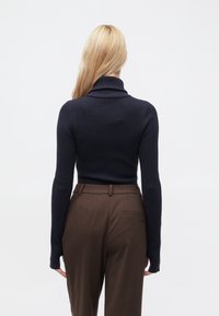 Maglione dolcevita a coste blu navy con maniche lunghe, abbinato a pantaloni sartoriali marrone scuro con tasca posteriore. Sfondo neutro.