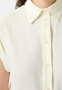 Blusa a maniche corte di colore giallo chiaro con bottone anteriore, colletto classico e tessuto morbido e strutturato con piccoli bottoni.