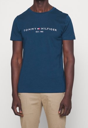 Mężczyzna ubrany w niebieską koszulkę Tommy Hilfiger z logo i napisem "EST. 1985", w beżowych spodniach, stojący na jednolitym tle.