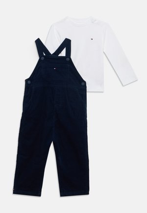 Salopette in velluto a coste blu navy con tasca frontale abbinata a una camicia bianca a maniche lunghe, entrambe con piccolo ricamo del logo.