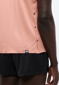 Αθλητική μπλούζα σε ροδακινί απόχρωση με υφή, με μικρές μαύρες κουμπότρυπες στα πλαϊνά και μαύρη ετικέτα λογότυπου κοντά στο τελείωμα.
