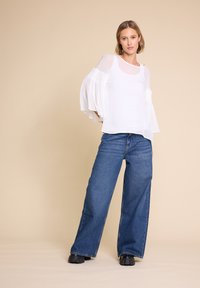 Hvit bluse med gjennomsiktige, volangermede ermer som er kombinert med løstsittende blå jeans. Blusen har en rund halslinje og en glatt tekstur.