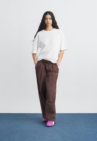 Νεαρή γυναίκα που στέκεται με τα χέρια στις τσέπες, φορώντας λευκό φαρδύ t-shirt, καφέ αθλητικό παντελόνι και ροζ αθλητικά παπούτσια Puma πάνω σε μπλε πάτωμα.