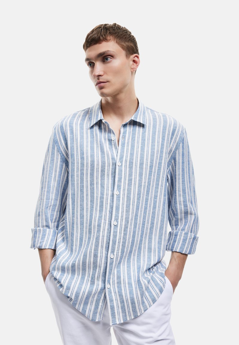 Vaaleansininen ja valkoinen raidallinen pitkähihaista paitaa, jossa on napillinen kaulus, valmistettu teksturoidusta kankaasta. Paitassa on käärityt hihat ja pyöristetty helma.