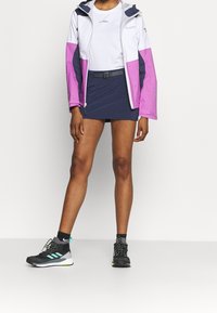 Chaqueta ligera en blanco, rosa y azul marino con capucha; combinada con un skort azul marino y zapatillas deportivas negras con detalles y cordones en verde azulado.