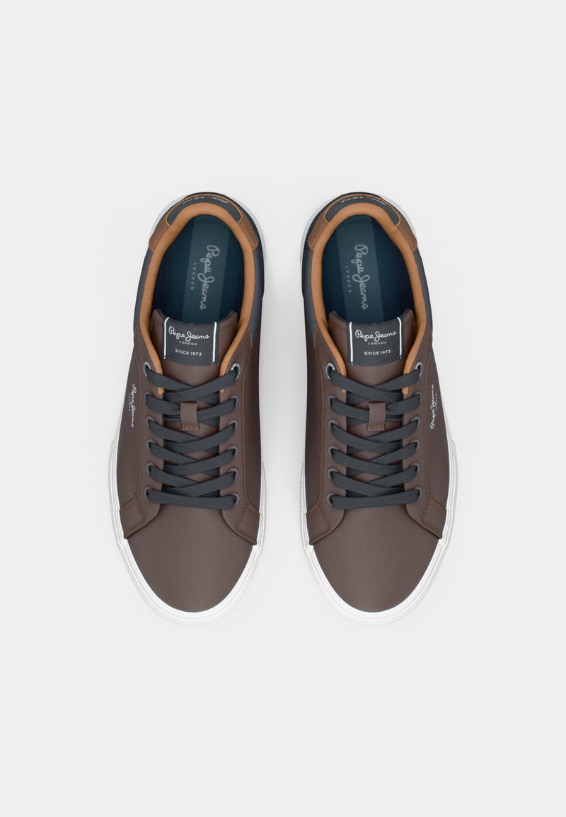 Zapatillas sintéticas marrones con detalles en azul marino, suelas de goma blanca y cordones negros. Presentan una etiqueta de logo en la lengüeta y en el costado.