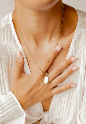 Gold necklace with an oval mother-of-pearl pendant, worn on bare skin. Soft white textured fabric visible in background. Neutral nail polish.