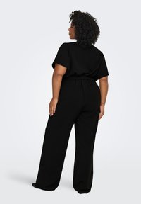 Juodas kombinezonas su trumpomis rankovėmis, juosmens diržu ir plačiomis kelnėmis. Pagamintas iš minkštos medžiagos, pasižymi laisvu kirpimu ir lygiu paviršiumi.