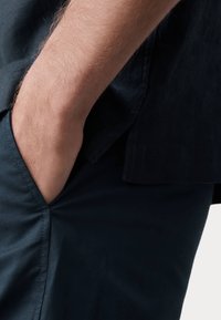 Pantalones azul oscuro con una textura suave, que presentan un bolsillo lateral. Una mano reposa en el bolsillo, mostrando la caída de la tela.
