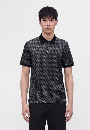 Ung mand stående med fronten mod kameraet iført en sort og hvid mønstret kortærmet polo shirt og sorte bukser mod en ensfarvet baggrund.