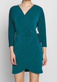 Teal omslagsklänning med djup V-ringning, trekvartsärmar och knytning i midjan. Tillverkad av mjukt, slätt tyg med en draperad siluett.