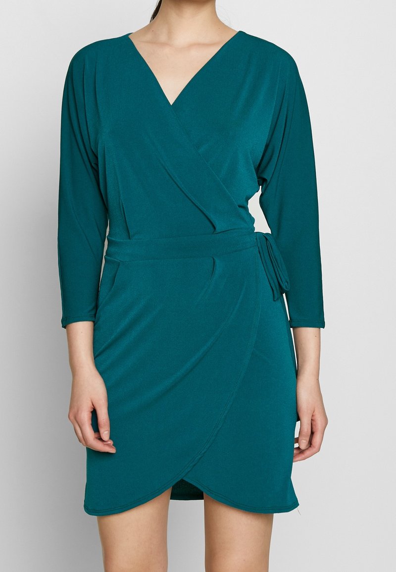 Teal omslagsklänning med djup V-ringning, trekvartsärmar och knytning i midjan. Tillverkad av mjukt, slätt tyg med en draperad siluett.
