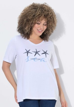 Frau mit lockigem Haar, die ein weißes T-Shirt mit drei Seesternen und dem Text „Ich brauche Vitamin See“ trägt, lächelnd und nach unten schauend, vor einer weißen Wand stehend.