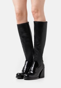Bottes noires montantes en matériau synthétique brillant, présentant une semelle plateforme, un talon épais et un détail de bride à la cheville. Fermeture zippée sur le côté.
