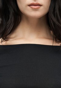 Κοντινό πλάνο του κάτω μέρους του προσώπου μιας γυναίκας, του λαιμού και των κλείδων της, που φοράει μαύρο τοπ χωρίς ώμους και έχει σκούρα καστανά μαλλιά.