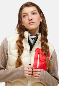 Gilet rembourré crème avec une poche poitrine zippée rouge. Il présente un col haut, un dessous texturé beige à manches longues et des tresses dans les cheveux.