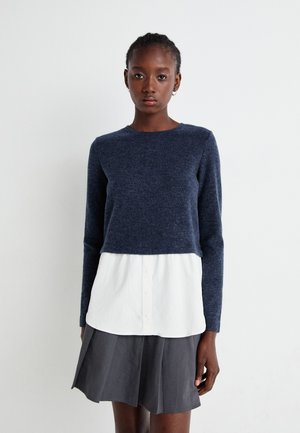 Pull en laine de couleur marine avec des manches longues, porté par-dessus une chemise blanche à boutons. La tenue est associée à une jupe plissée grise.