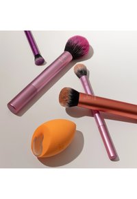 Vijf make-upkwasten met roze en koperen handvatten en een oranje make-upspons met foundationresten op een witte ondergrond.