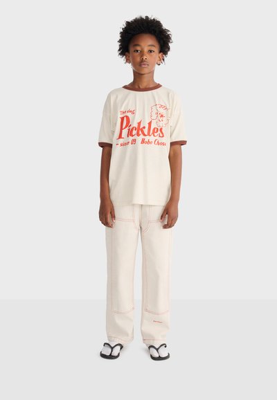 Child stands facing forward wearing cream T-shirt with red text "Pickles" and matching cream pants with visible red stitching and black sandals.