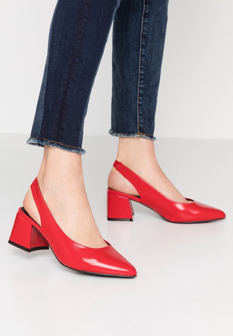 Personne portant un jean bleu foncé à ourlet effiloché et des chaussures rouges à talons bas à bride arrière sur un fond de sol clair.