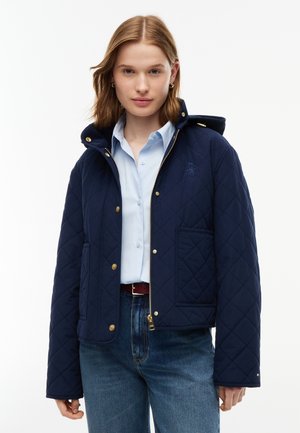 Ung kvinde iført en mørkeblå quiltet jakke med guldknapper over en lyseblå skjorte og blå jeans, vendt fremad med et neutralt udtryk.