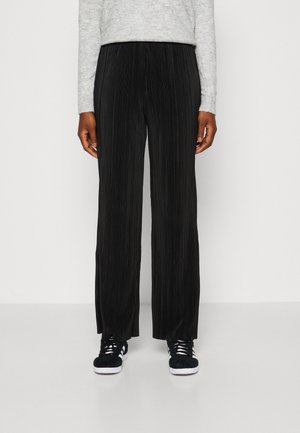 Person trägt schwarze, weit geschnittene, plissierte Hose, grauen Strickpullover und schwarze Turnschuhe mit weißen Sohlen und Schnürsenkeln, steht auf einem schlichten Boden.