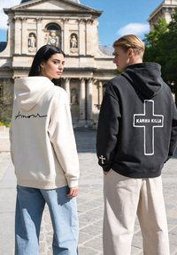 Deux jeunes personnes sont dehors près d’un bâtiment historique, portant des sweats à capuche avec les inscriptions « Amour » et « Karma Kills » dans le dos.
