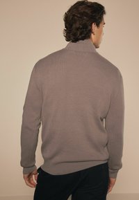 Grå strikket pullover med høj krave, ribbede manchetter og lige kant, båret med sorte bukser, set fra ryggen.