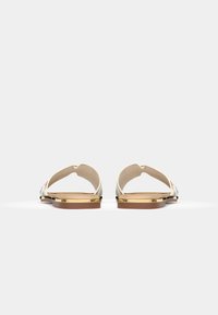 Sandales à bout ouvert avec une lanière à motifs, accents dorés métalliques et une semelle marron clair. Texture lisse et design minimaliste.