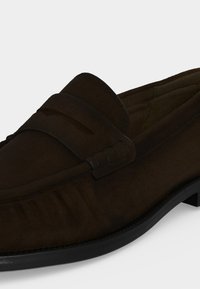 Mocassin en daim marron foncé avec un petit talon noir, bout rond et un détail de sangle sur le dessus avant.