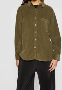 Oliwkowa koszula z koronką, zapinana na guziki, z kołnierzykiem i jedną kieszenią na piersi, w zestawie z czarnymi spodniami w prążki.