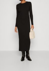 Svart långärmad klänning, figurnära design, halvlång längd. Bärs med svarta klackstövlar och en liten beige plyschhandväska. Slät textur.