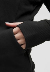 Black sweatshirt featuring a ribbed cuff and a hand tucked into a front pocket. The fabric appears soft and slightly textured.