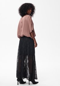 Vrouw met krullend haar die een losse mauve blouse, een lange zwarte kanten rok en zwarte heeled enkellaarsjes draagt, staand tegen een witte achtergrond.