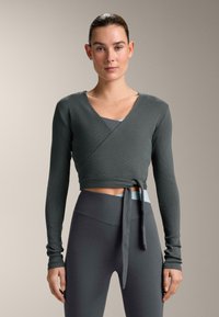 Top grigio a maniche lunghe con scollo incrociato e fiocco in vita, caratterizzato da una trama a coste. Indossato sopra una canotta sportiva azzurro chiaro e abbinato a leggings scuri.