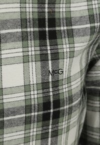 Groene en zwarte plaid stof met witte accenten; bevat een geweven textuur en N&G-logo in het zwart. Geaccentueerde vierkanten in verschillende groottes.