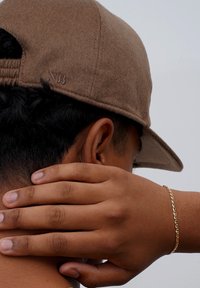 Braune Wollmütze mit flachem Schirm, featuring ein gesticktes Logodetail. Am Hals platziert, trägt einen goldenen Kettenarmreif.