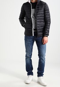 Svart pufferjacka med horisontella sömmar, ståkrage och vit dragkedja. Tillsammans med en randig svart och vit skjorta, blå jeans och vita sneakers.