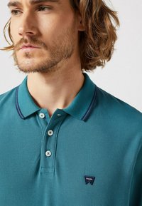 Turkos polo skjorta i texturerad tyg, tre-knapplåsning och kontrasterande marinblå kragstripes. Har en liten Wrangler-logotyp på bröstet.