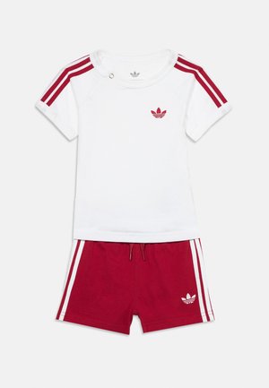 Chemise blanche à manches courtes avec des rayures rouges sur les épaules et un short rouge assorti avec des rayures blanches, tous deux avec le logo Adidas.