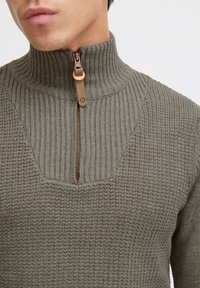 Olivgrüner Strickpullover mit einem Reißverschluss-Hoheitskragen, ausgestattet mit gerippter Struktur und einem einzigartigen V-förmigen Ausschnitt.
