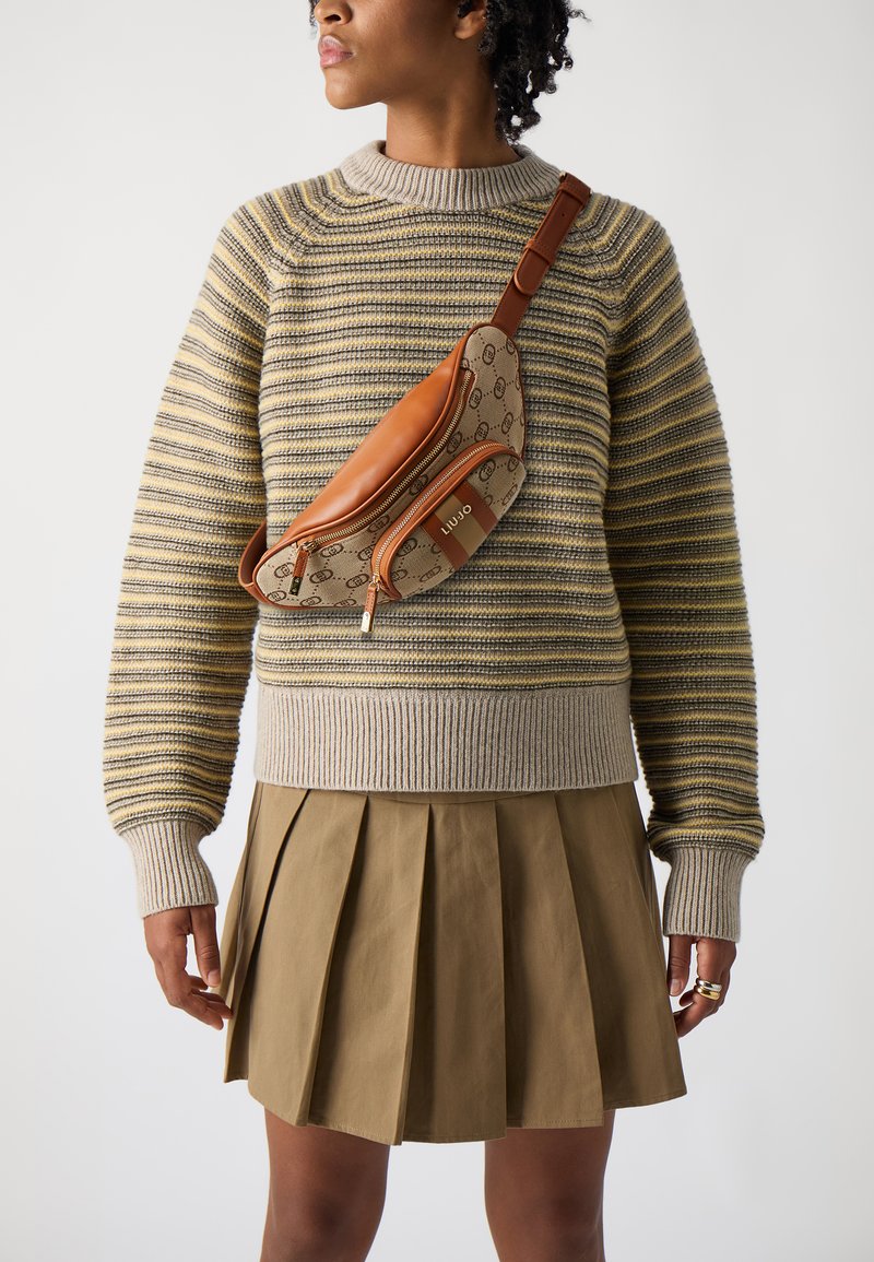 Braun und beige gemusterte Gürteltasche mit Reißverschluss und Logo, getragen über einem grau und gelb gestreiften Pullover, kombiniert mit einem khakifarbenen Plisseerock.