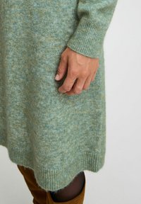 Abito in maglia realizzato in un tessuto morbido e strutturato, caratterizzato da un colore verde tenue, polsini a coste e un orlo scallop. Mano visibile sul lato.