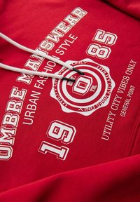 Sweat à capuche rouge en tissu doux avec impression graphique blanche, comportant le texte "OMBRE MANSWEAR", des motifs circulaires et des cordons de serrage avec embouts noirs.