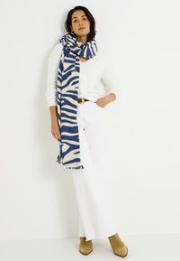 Écharpe à motif zébré en mélange de laine bleu marine et crème, avec des bords à franges. Portée sur un pull blanc et associée à un jean évasé et des bottes.