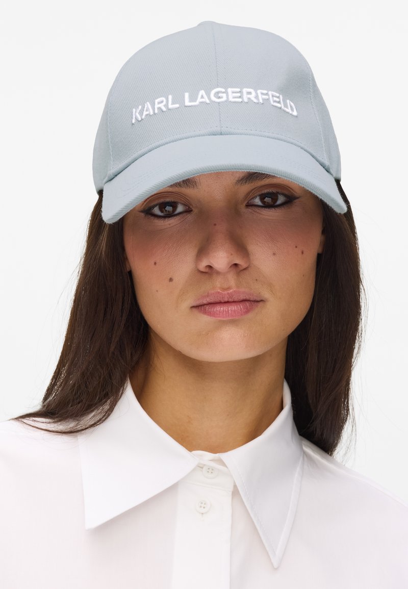 Γυναίκα που φορεί ανοιχτό μπλε καπέλο μπέιζμπολ με το κείμενο "KARL LAGERFELD" και λευκό πουκάμισο με γιακά, κοιτώντας κατευθείαν μπροστά.