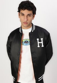 Jachetă bomber neagră din satin cu guler și manșete albe stripate, având un logo alb cu litera 'H'. Sub aceasta, o tricou alb cu o grafică colorată.